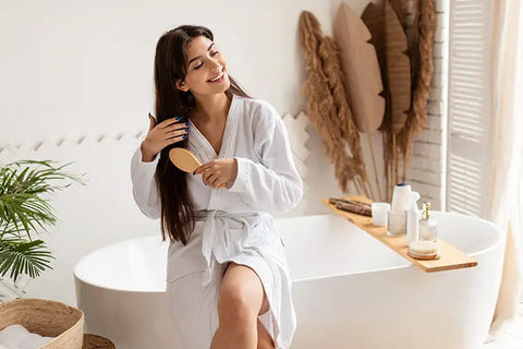brunette woman sitting on the side of bathtub brushing long hair extensions