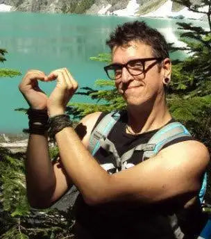 A person wearing black-framed glasses and a backpack, making a heart shape with their hands.
